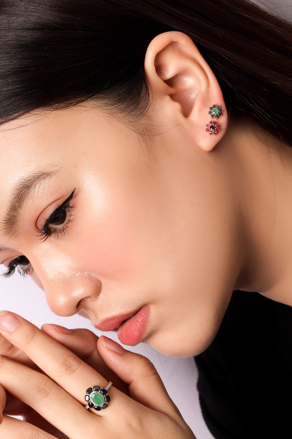 Close-up of a woman wearing a silver ring with a green and black floral design on her finger, along with matching stud earrings in her ear, against a light background.