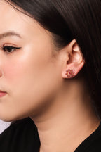 A close-up side profile of a woman wearing a small floral stud earring in pinkish-red and gold-toned metal.
