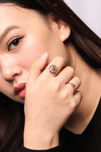 A close-up lifestyle shot of a woman wearing a gold ring with a green and pink gemstone cluster on her finger, with a second gold ring featuring pink gemstones on her middle finger.