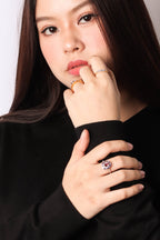 A close-up of a woman wearing a prominent ring with a large pinkish gemstone and smaller clear stones on a gold band, styled with other rings on her fingers, set against a plain light background.
