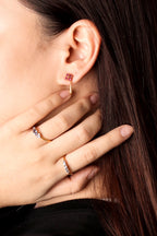 A close-up of a woman's ear and hand wearing a delicate gold huggie hoop earring with a small red gemstone at the top, along with two gold rings on her fingers.