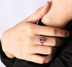 A close-up of a person's hand wearing a silver ring with a central round pink gemstone surrounded by small clear stones, set against a black top and light gray background.