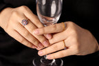 Close-up of a person's hands holding a clear wine glass against a black background, wearing multiple rings including a prominent pink and blue flower-shaped ring, a gold band with a blue stone, and a gold band with a small blue stone.