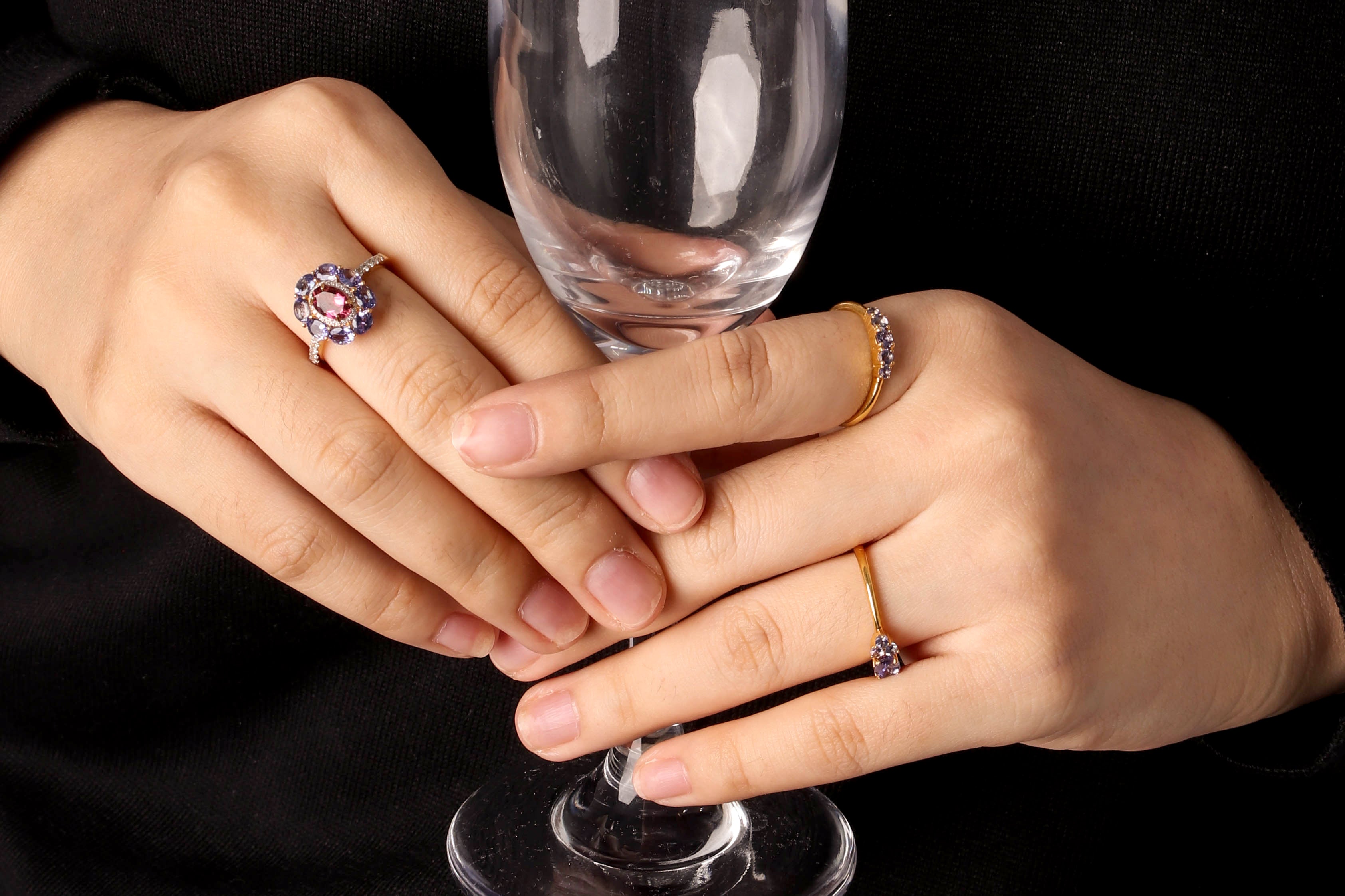 Close-up of a person's hands holding a clear wine glass against a black background, wearing multiple rings including a prominent pink and blue flower-shaped ring, a gold band with a blue stone, and a gold band with a small blue stone.