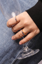 A close-up of a person's hand wearing two delicate gold rings with small dark blue gemstone accents, resting on blue denim fabric next to a clear glass.