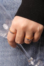 A close-up of a hand wearing two gold rings with small clear gemstones, resting on blue denim next to a clear glass.