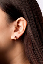 A close-up side profile of a woman's ear wearing a small rose gold stud earring with a dark red gemstone.