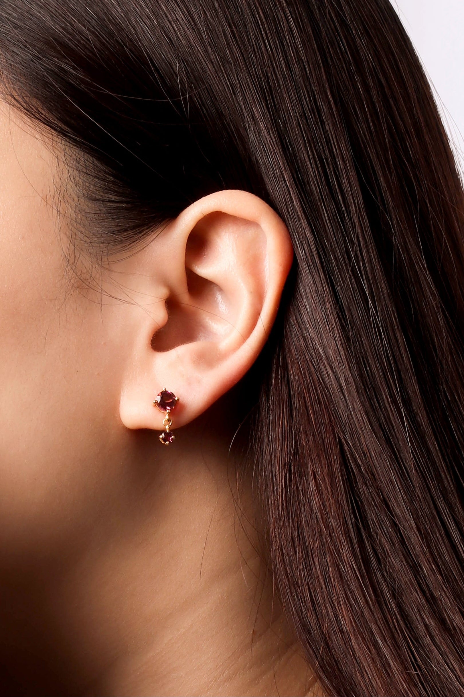 A close-up side profile of a woman's ear wearing a small rose gold stud earring with a dark red gemstone.
