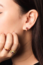 Close-up of a woman's ear wearing a small gold stud earring with a pink gemstone, with her hand resting near her face showing matching gold rings.