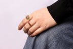 A close-up of a person's hand resting on their lap, wearing two gold rings: one with a round green and pink stone, and another with a curved row of small pink stones.