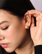 A close-up of a woman wearing a small gold hoop earring with a dangling heart-shaped charm, holding her ear to show the fit.