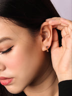 A close-up of a woman wearing a small gold hoop earring with a dangling heart-shaped charm, holding her ear to show the fit.