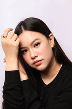 A close-up portrait of a woman with long dark hair, wearing a black long-sleeve top and small gold hoop earrings, posed against a light gray studio background.