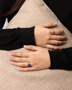 A close-up of a person's hands resting on a beige woven cushion, wearing a prominent silver ring with a large oval blue gemstone and a smaller gold ring on the other hand.