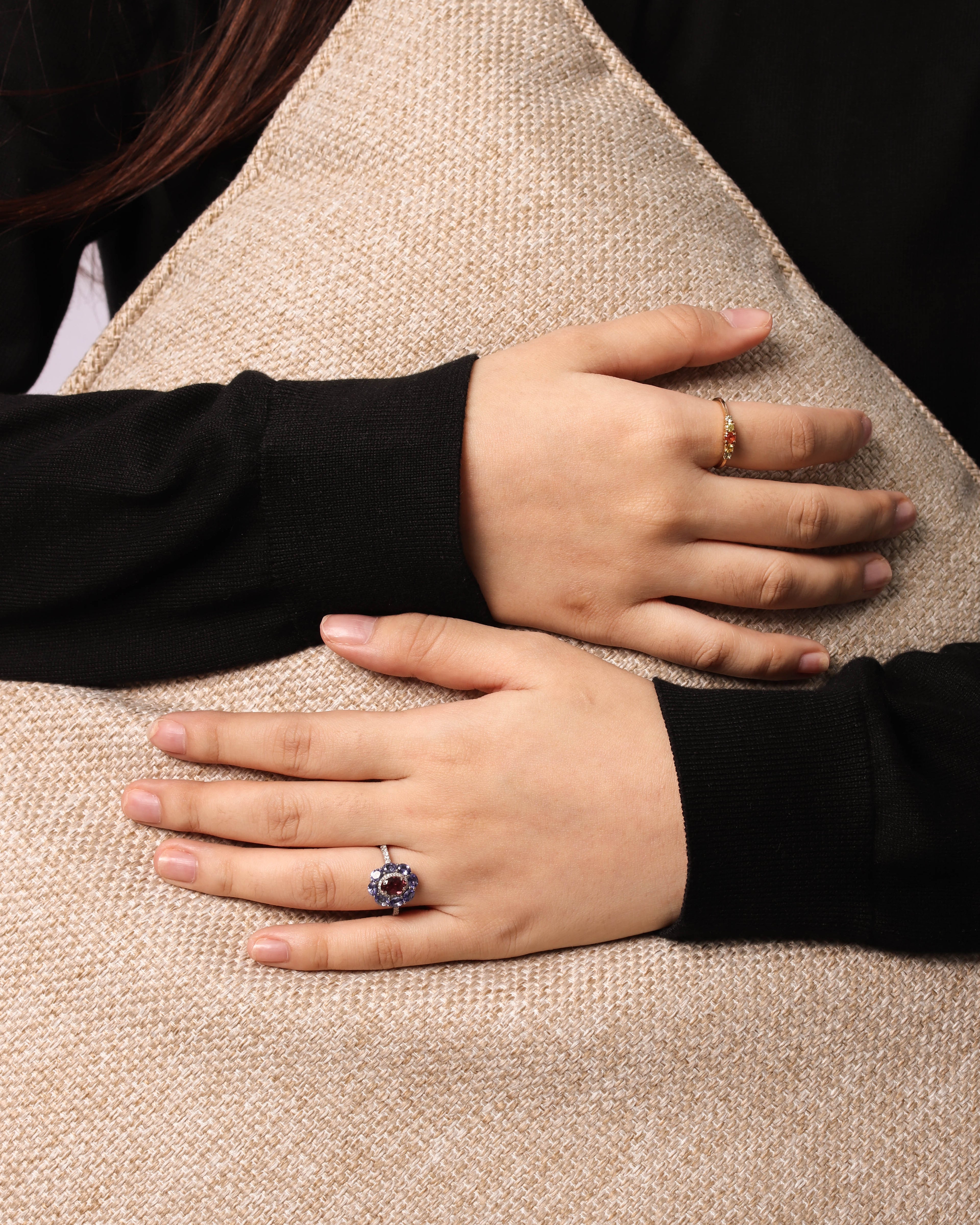 A close-up of a person's hands resting on a beige woven cushion, wearing a prominent silver ring with a large oval blue gemstone and a smaller gold ring on the other hand.