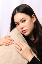 A woman with long dark hair is shown in a close-up portrait, wearing a ring with a large oval blue gemstone and a gold band, while resting her head on a textured beige pillow.
