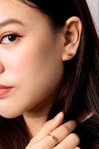 Close-up of a woman wearing a small gold stud earring with a red and yellow stone cluster, along with a gold ring on her finger.