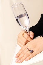 A close-up of a person's hands resting on a white surface, with one hand wearing a gold band and the other wearing a ring with a purple and blue gemstone.