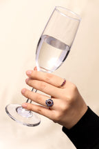 A hand holding a clear wine glass against a light beige background, wearing a silver ring with a dark purple and blue floral center and a smaller matching ring on the other finger.