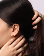 A close-up of a woman with dark hair wearing a small gold earring with a dangling teardrop-shaped charm.