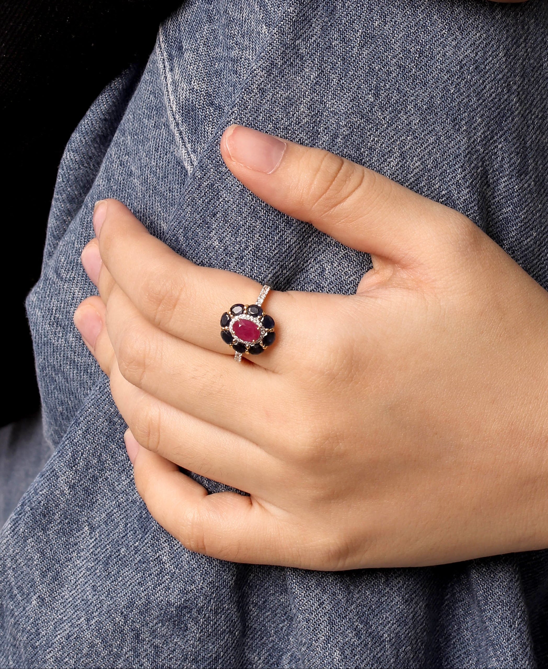 Close-up of a person's hands wearing multiple rings, including a prominent ring with a dark red flower-shaped stone and black petals, a green rectangular stone ring, and gold bands, set against a black top and blue denim.