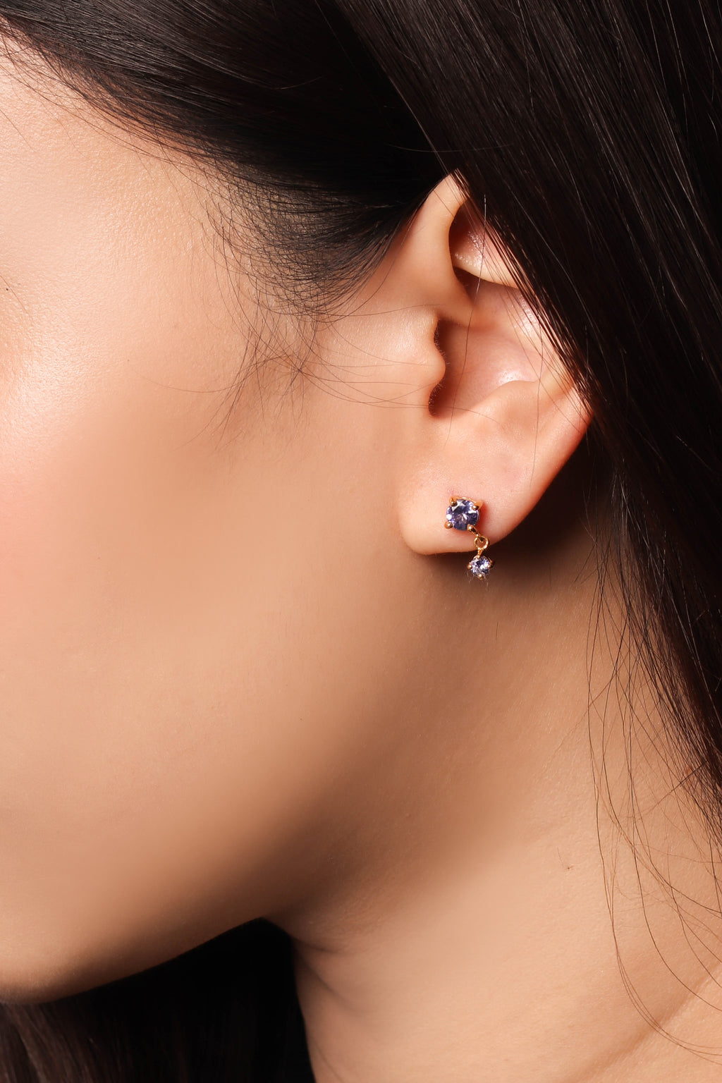 A close-up of a person's ear wearing a small gold huggie hoop earring with a cluster of deep blue gemstones and a dangling clear crystal.