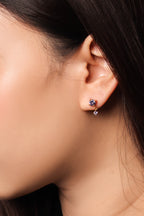 A close-up of a person's ear wearing a small gold huggie hoop earring with a cluster of deep blue gemstones and a dangling clear crystal.