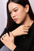 A close-up portrait of a woman wearing delicate gold jewelry, including a thin gold chain necklace with a dark bead pendant, a gold ring on her middle finger, and a gold ring with a green stone on her ring finger, against a soft light gray background.