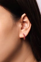 A close-up of a model's ear wearing a small curved hoop earring set with vibrant pink gemstones.