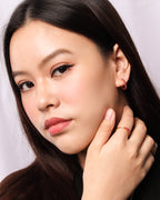 A close-up portrait of a woman with long dark hair wearing a small gold hoop earring with a red gemstone and a matching gold ring with a red gemstone.