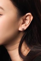 A close-up side profile of a woman wearing a small floral stud earring in dark gold with green and pink stones.