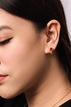 A close-up of a woman's ear wearing a small gold stud earring with a dark red gemstone, against a light neutral background.