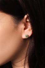 A close-up of a person's ear wearing a small circular stud earring with green and red stones, set against dark hair and a dark background.