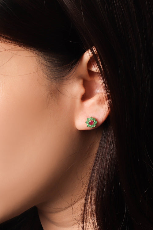 A close-up of a person's ear wearing a small circular stud earring with green and red stones, set against dark hair and a dark background.