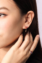 A close-up of a woman's ear wearing a small gold hoop earring with a red gemstone cluster, with her hand resting near her face. She is also wearing a gold ring with red and blue stones.
