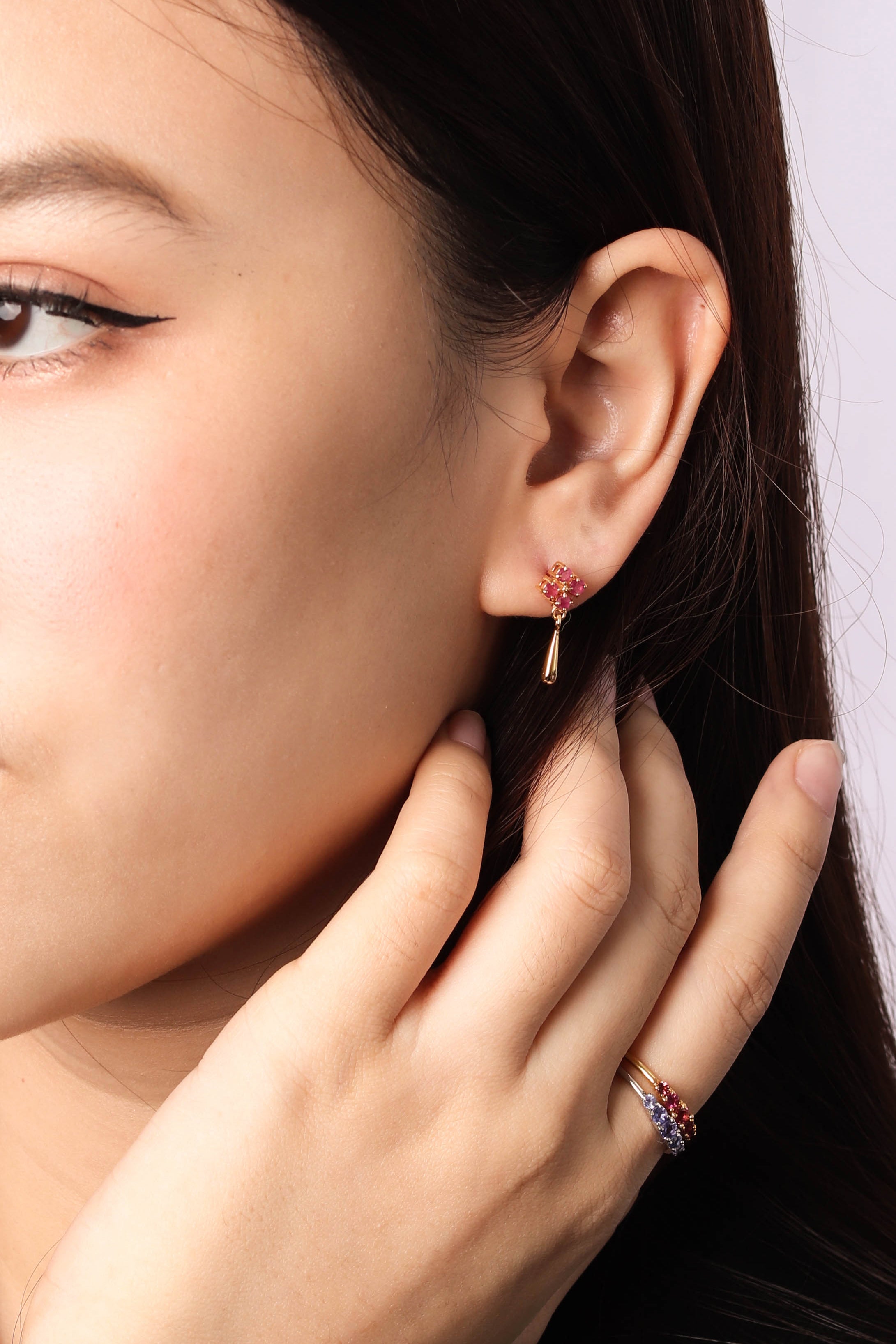 A close-up of a woman's ear wearing a small gold hoop earring with a red gemstone cluster, with her hand resting near her face. She is also wearing a gold ring with red and blue stones.
