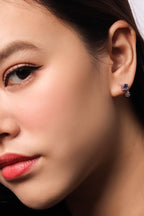 Close-up of a woman's ear wearing a small dangling stud earring with a dark blue stone and silver-toned metal.