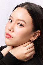 A close-up portrait of a woman with dark hair wearing a small gold hoop earring with a dark gemstone, posing with her hands near her neck against a light gray background.