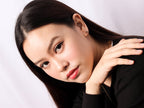A close-up portrait of a woman with long dark hair, wearing a black top and a small gold hoop earring, set against a plain light background.