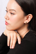 A close-up side profile of a woman wearing a small gold ring with green and red stones on her finger and a matching earring, wearing a black top against a light gray background.
