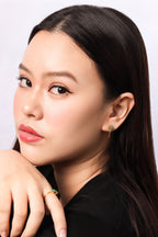 A close-up portrait of a woman with long dark hair, wearing a small gold ring with a green stone on her finger and a matching earring, set against a plain light gray background.