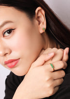 Close-up of a woman wearing a small gold ring with a green stone on her finger and a matching green-and-gold earring, against a soft light gray background.