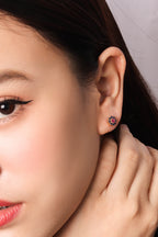 Close-up of a woman's ear wearing a small floral stud earring with pink and purple stones on a dark metal setting.