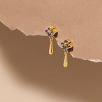 A pair of gold dangle earrings with blue gemstone floral clusters, displayed against a textured beige background with a torn-paper effect.
