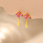 A pair of gold drop earrings with pink gemstone floral clusters and teardrop-shaped gold pendants, displayed against a soft beige textured background.