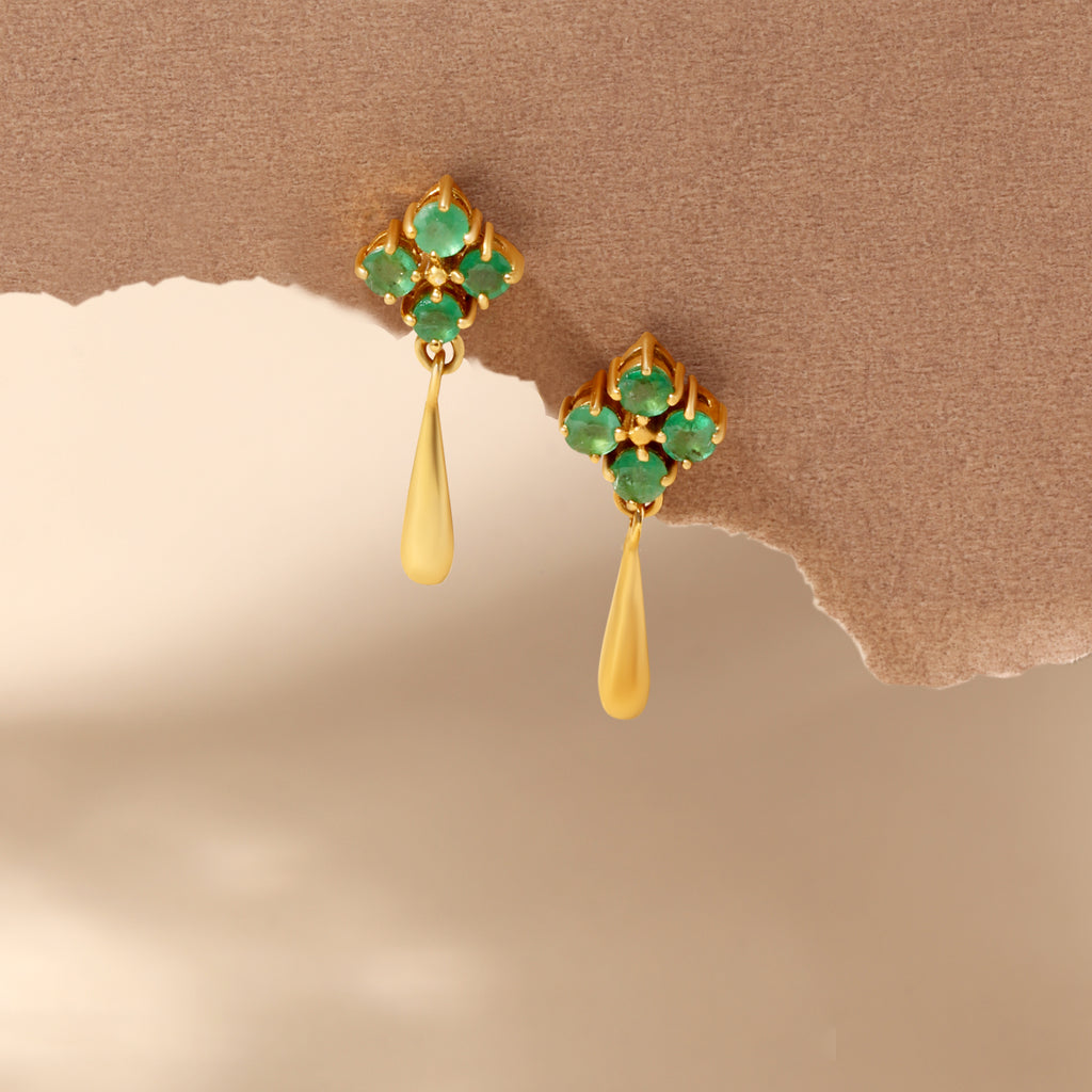 A pair of gold dangle earrings with green gemstone floral clusters and teardrop-shaped gold drops, displayed against a textured brown and beige background.