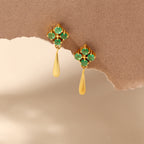 A pair of gold dangle earrings with green gemstone floral clusters and teardrop-shaped gold drops, displayed against a textured brown and beige background.