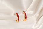 A pair of gold crescent-shaped hoop earrings set with small round red gemstones, displayed on draped white satin fabric.