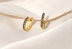 A pair of gold huggie hoop earrings embellished with small round blue gemstones, displayed on draped white satin fabric.