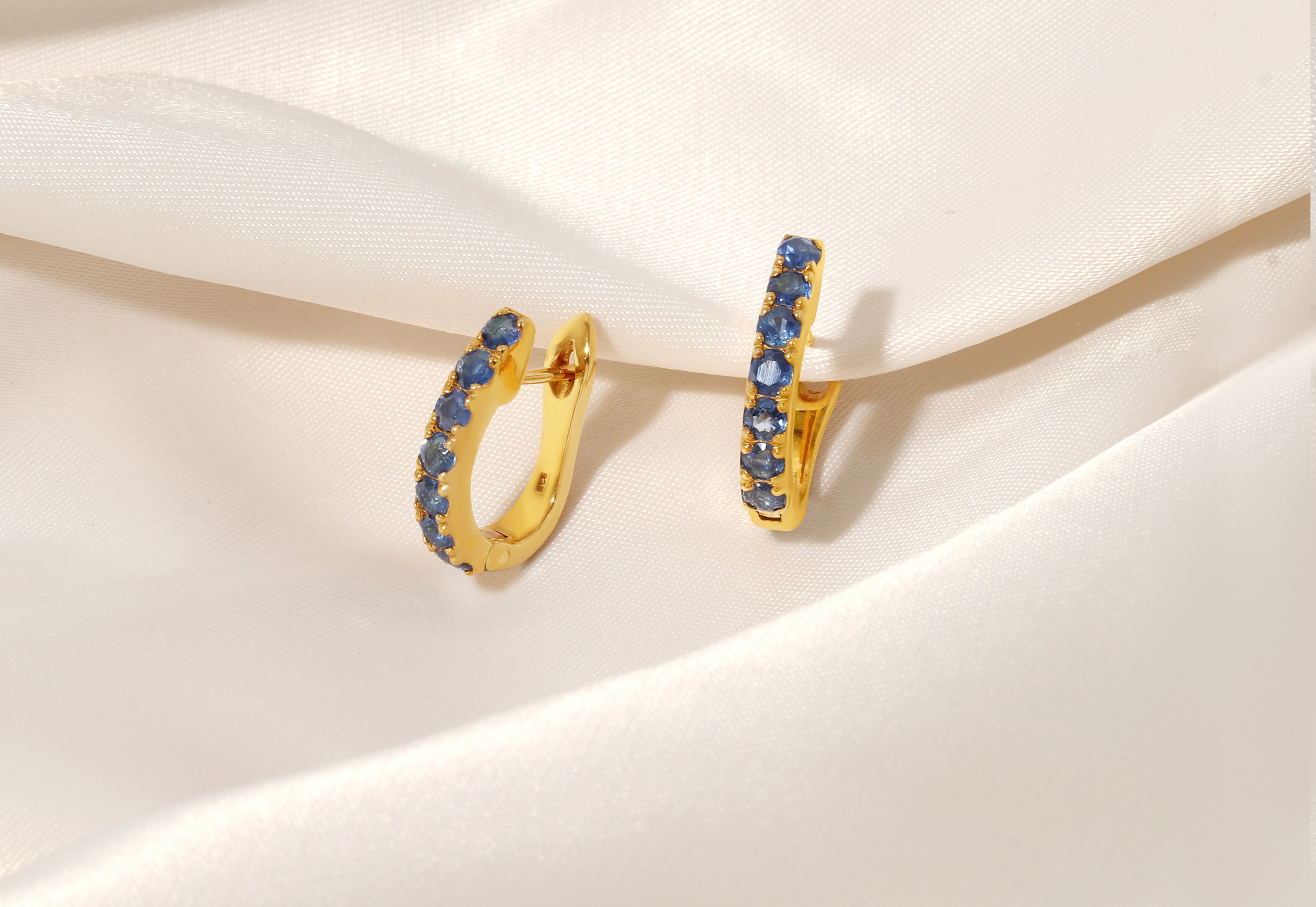 A pair of gold huggie hoop earrings embellished with small round blue gemstones, displayed on draped white satin fabric.
